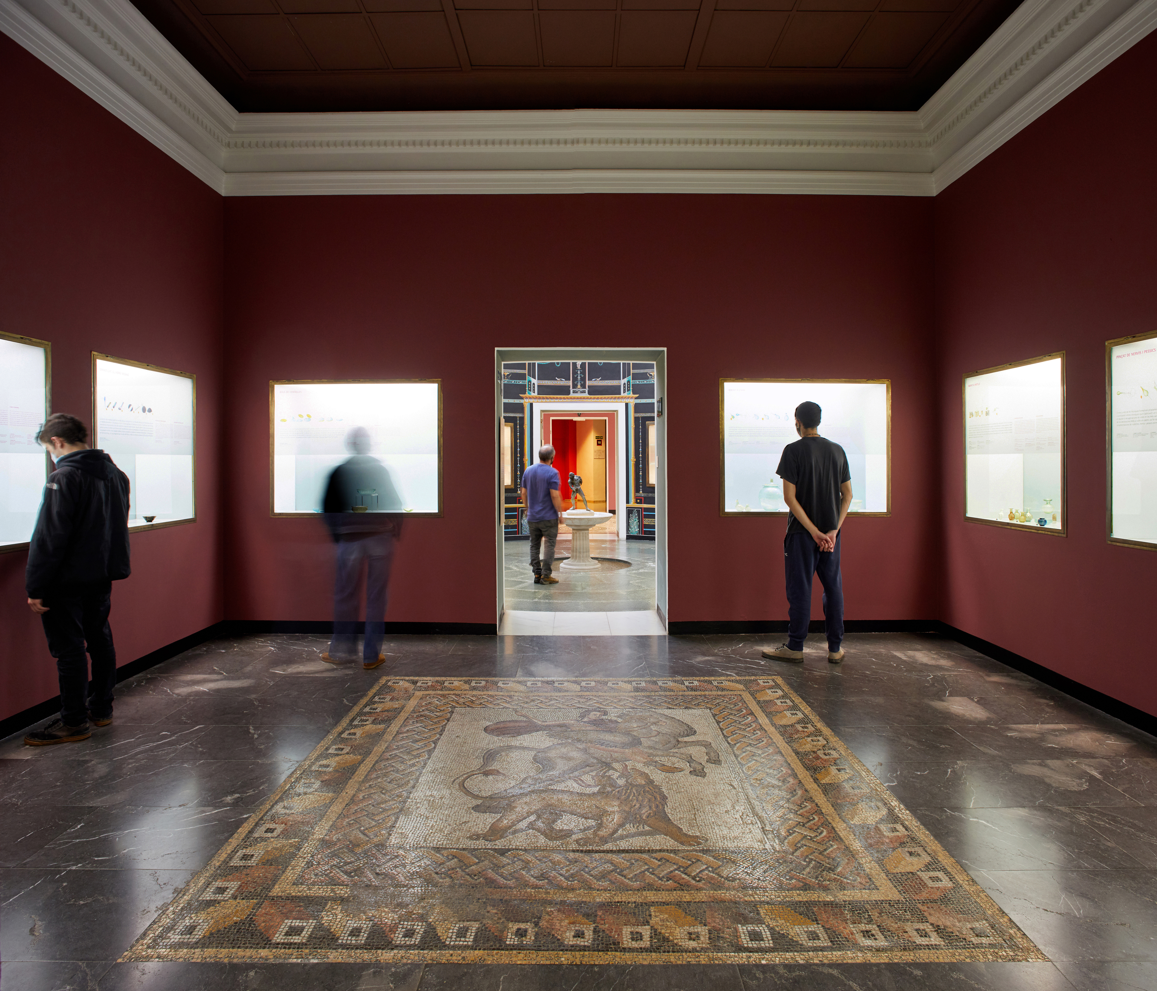 Interior d'una sala de museu amb mosaics al terra, vitrines i visitants.