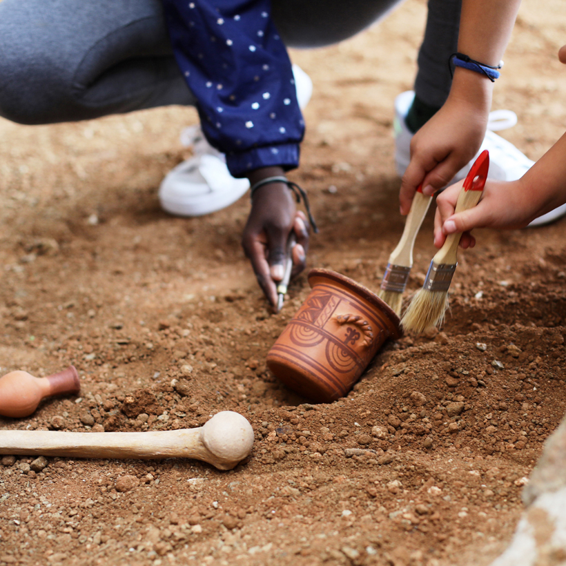 Mans d'infants agafant pinzells per desenterrar peces arqueològiques. Al mig de la imatge hi apareix un vas de ceràmica i un os.